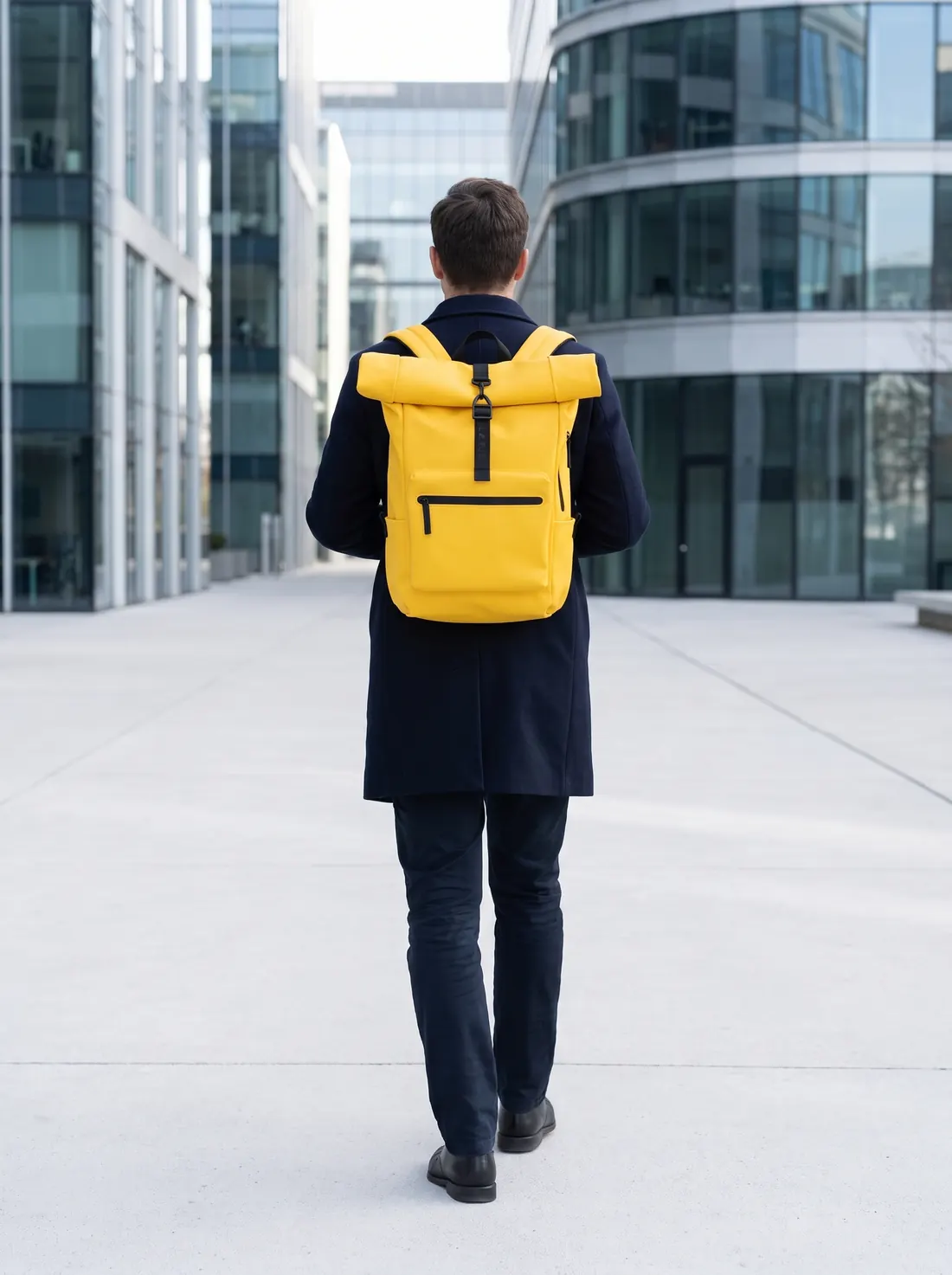 A person with their back turned crosses an office plaza with a yellow Pilatus Urban backpack